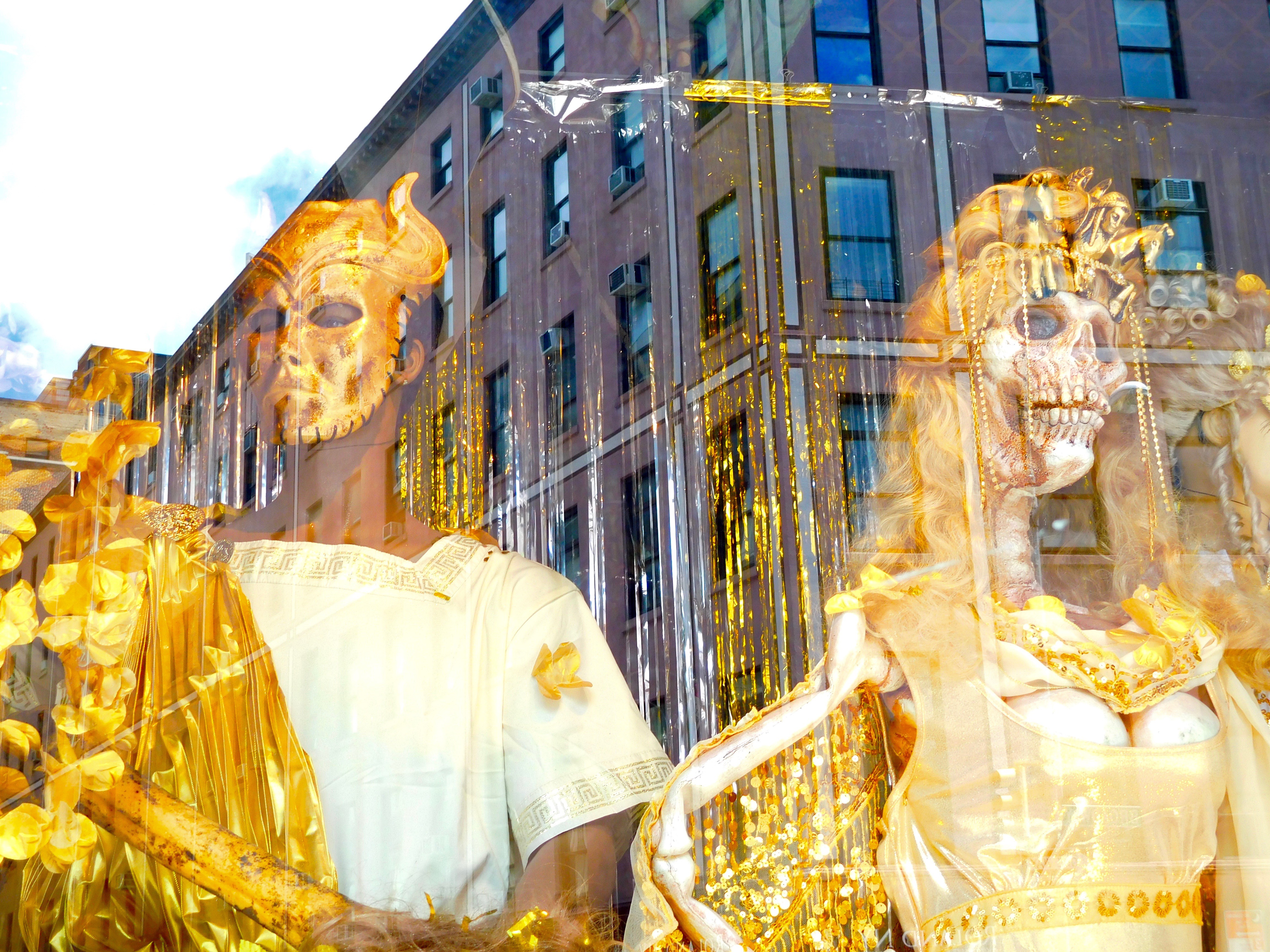 Two golden figures, one a skeleton, are displayed in a window that shows a reflection of the building behind it.
