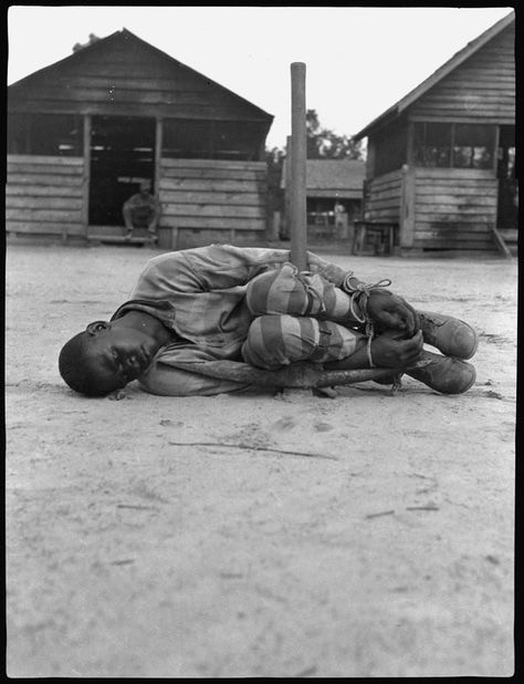 Photograph of young black boy shackled to pick with man in background looking on, seated on a porch.