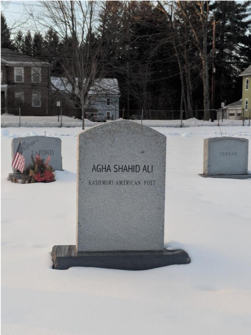 A gravestone that reads "Agha Shahid Ali" and under it "Kashmiri-American Poet" in the foreground with other headstones and houses in the background. The ground is covered in snow.