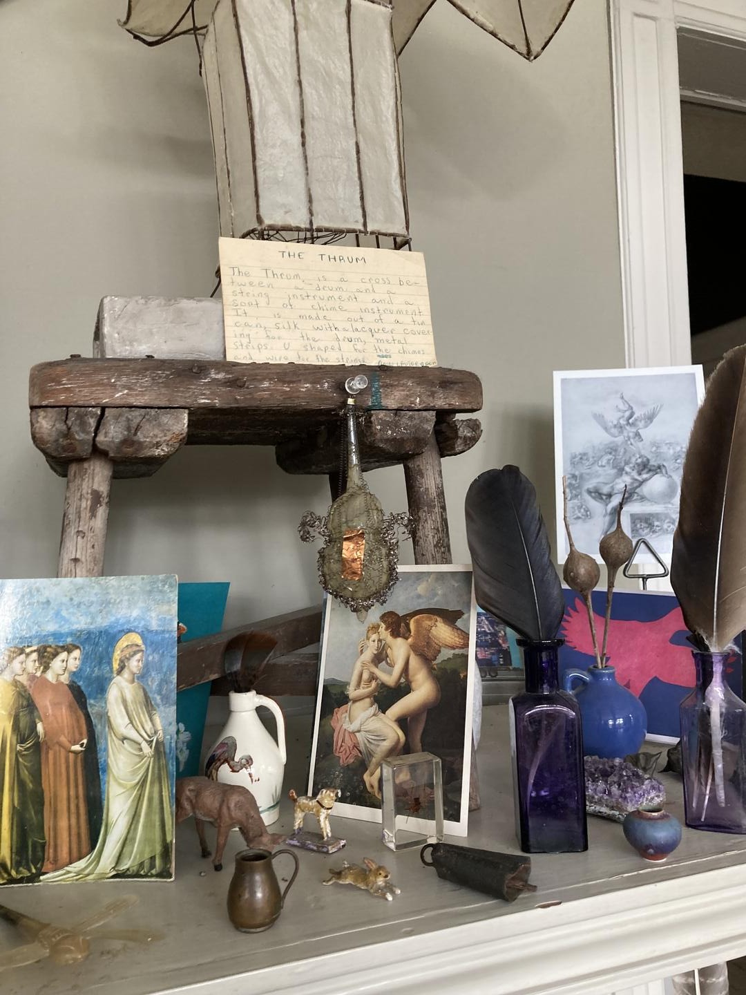 Color photo: Various items on a mantle including art prints, feathers, and figurines