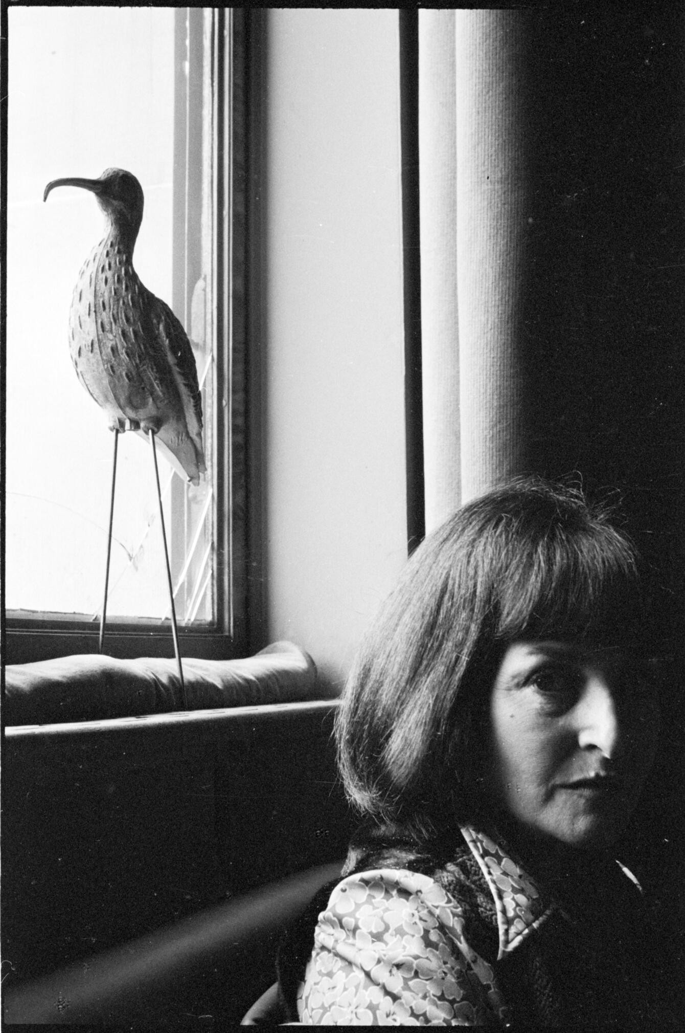 Black and white photo of a woman in profile posing in front of a window. Bright light casts shadows on her face. A bird sculpture is behind her.