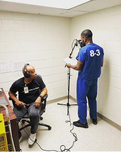 Two men in a small room that is painted white. One is standing at a microphone looking down at a sheet of paper in his hands. The other is sitting at a desk wearing headphones and looking at the other man.