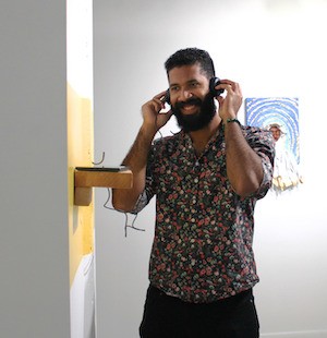 Smiling man with a beard wearing a flowered shirt in an art gallery listening to a poem on headphones near the poem's installation of a tablet on a small wooden shelf with a yellow accent wall.