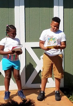  Two kids standing in front of a green wooden structure holding books that they appear to be speaking from.