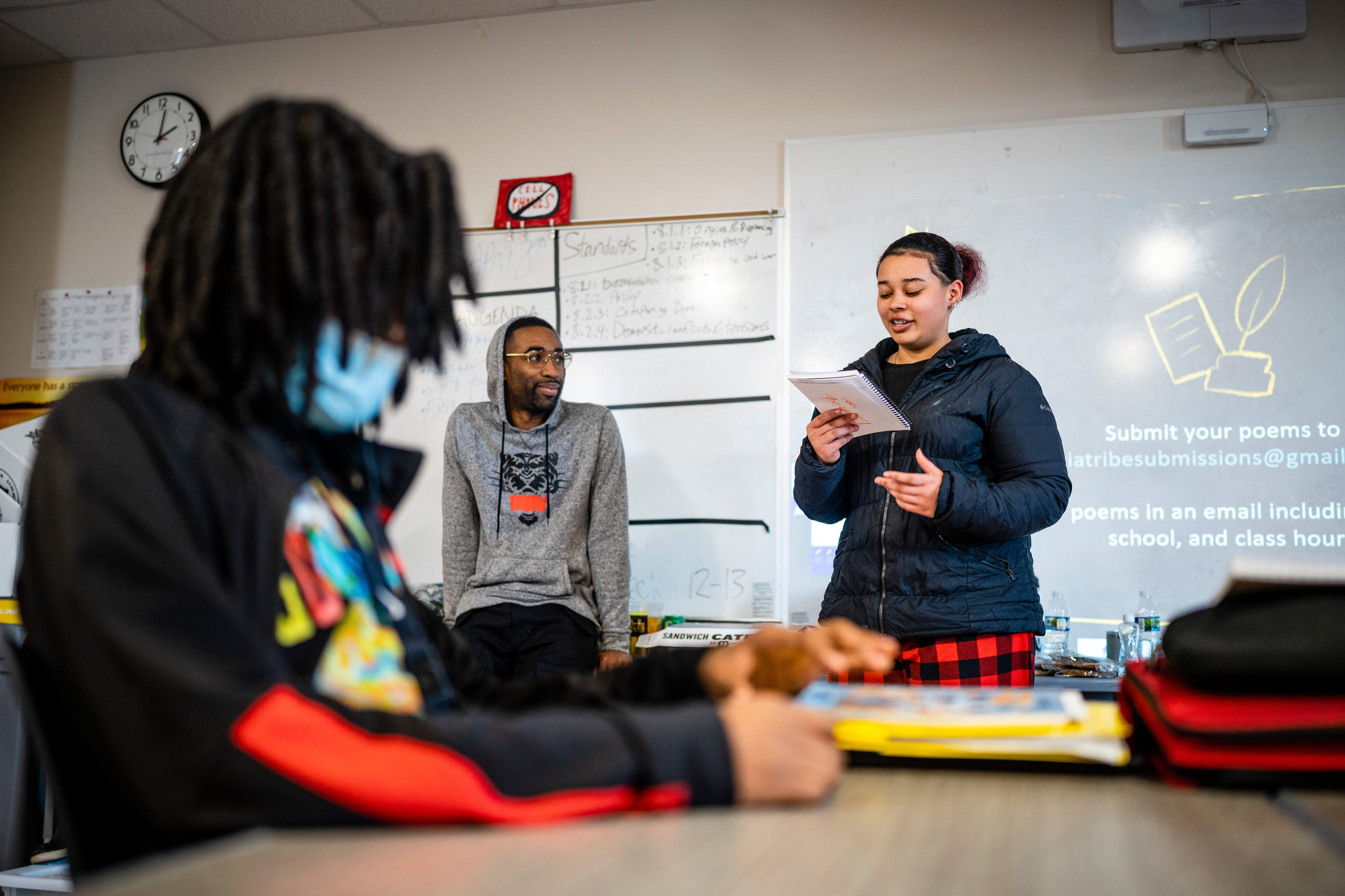 A young student smirking, with light brown skin and dark red hair reading their spoken word poetry in the classroom next to teaching artist Caleb Rainey.