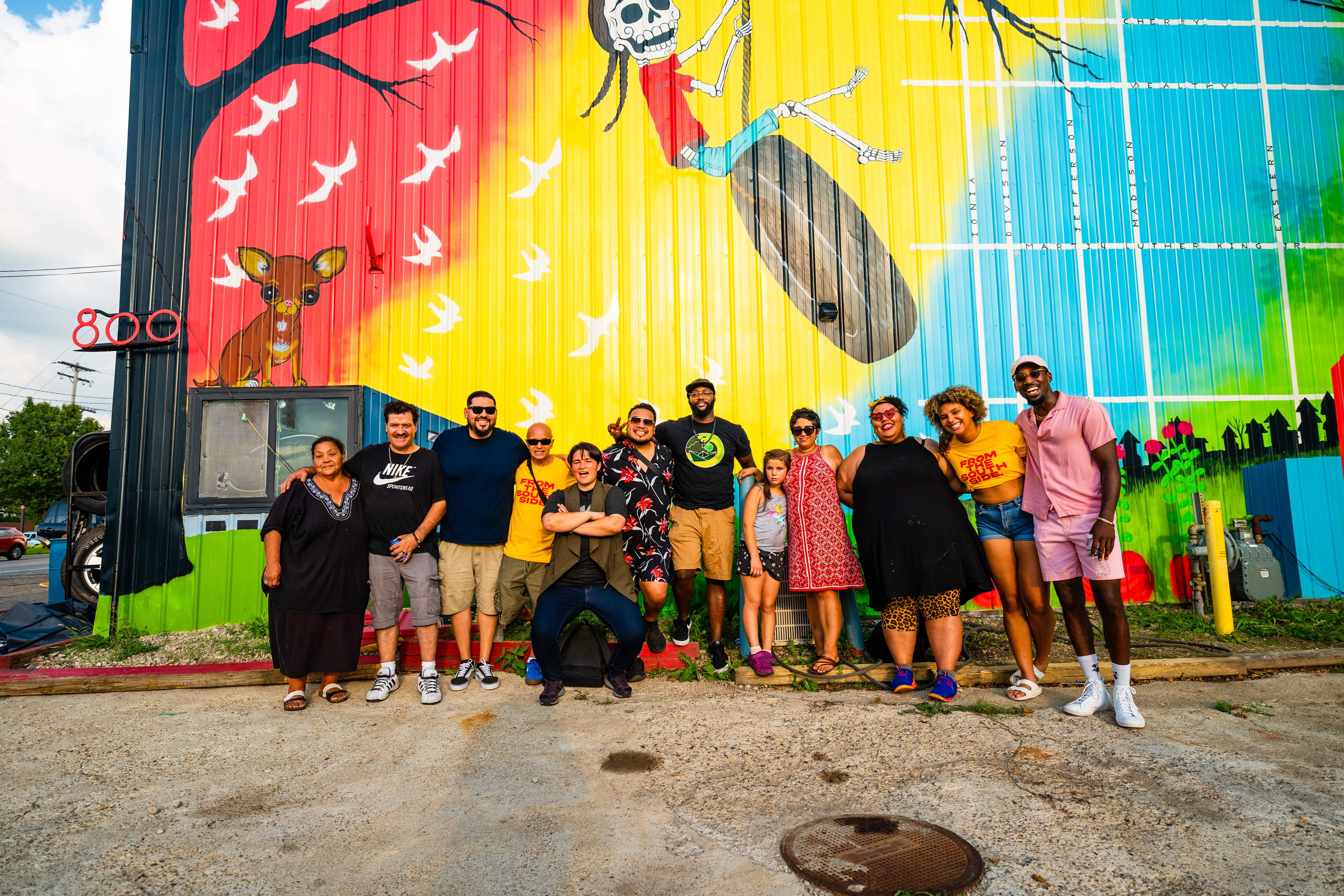 A group of Black and Brown folks and young people standing in front of a colorful mural done by local artist Alynn Guerra of Red Hydrant Press.