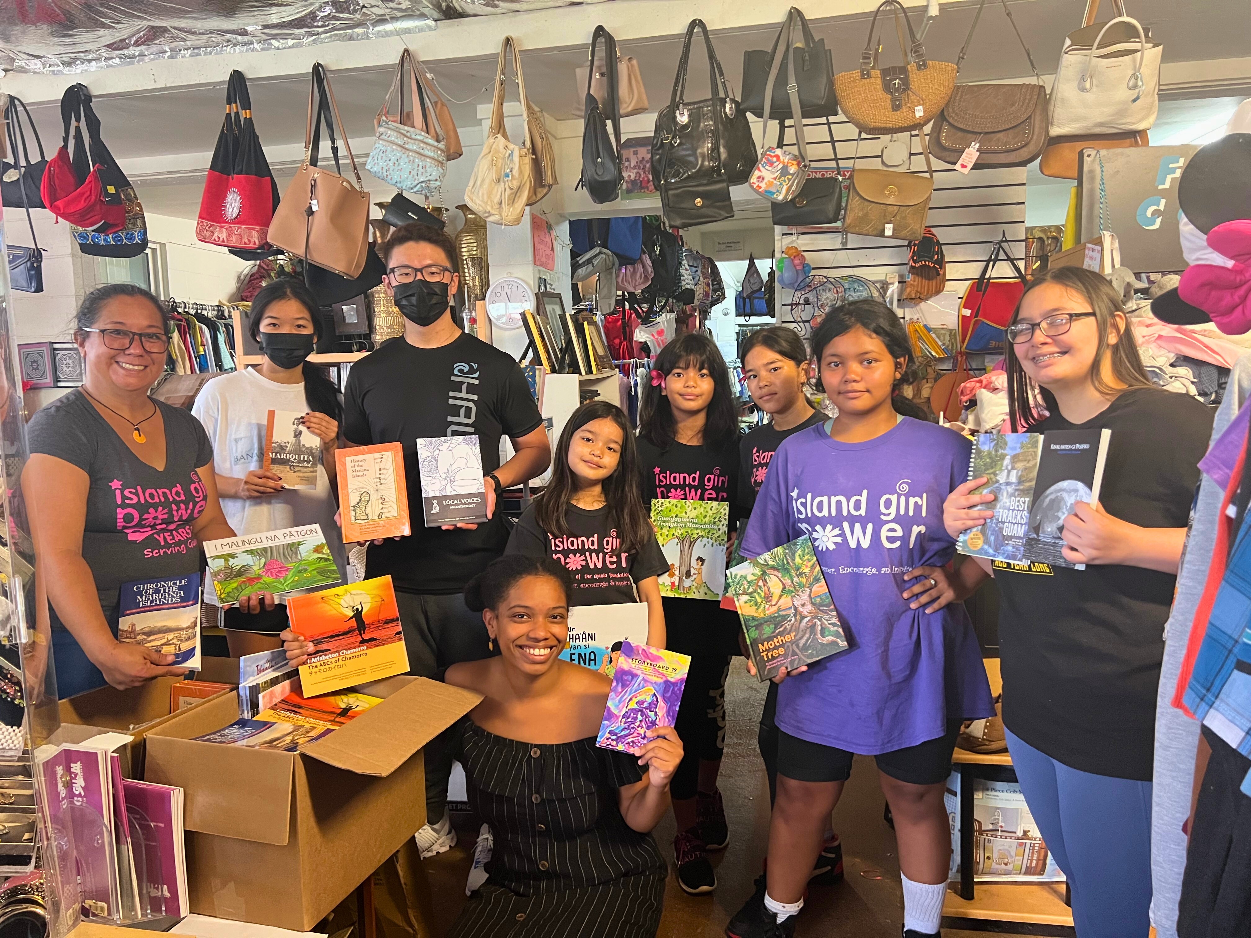 Group of young adults and kids holding up books inside a store with handbags hanging from the ceiling.