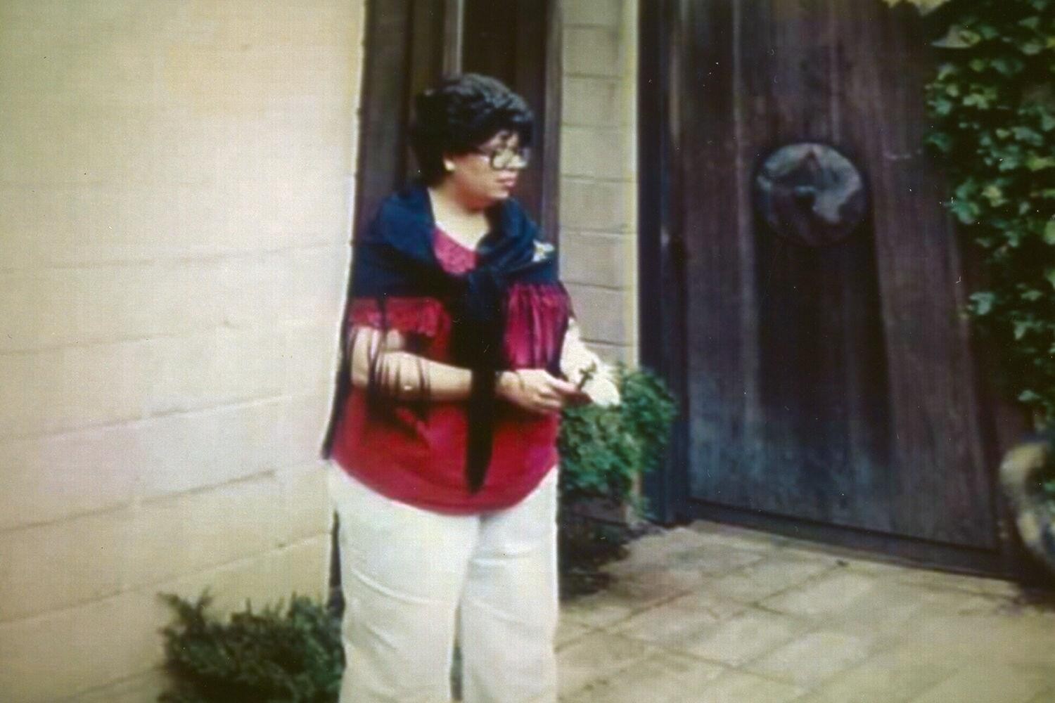 A photograph of a woman with short, dark hair and glasses wearing a red top, blue scarf, and white pants in a courtyard garden.