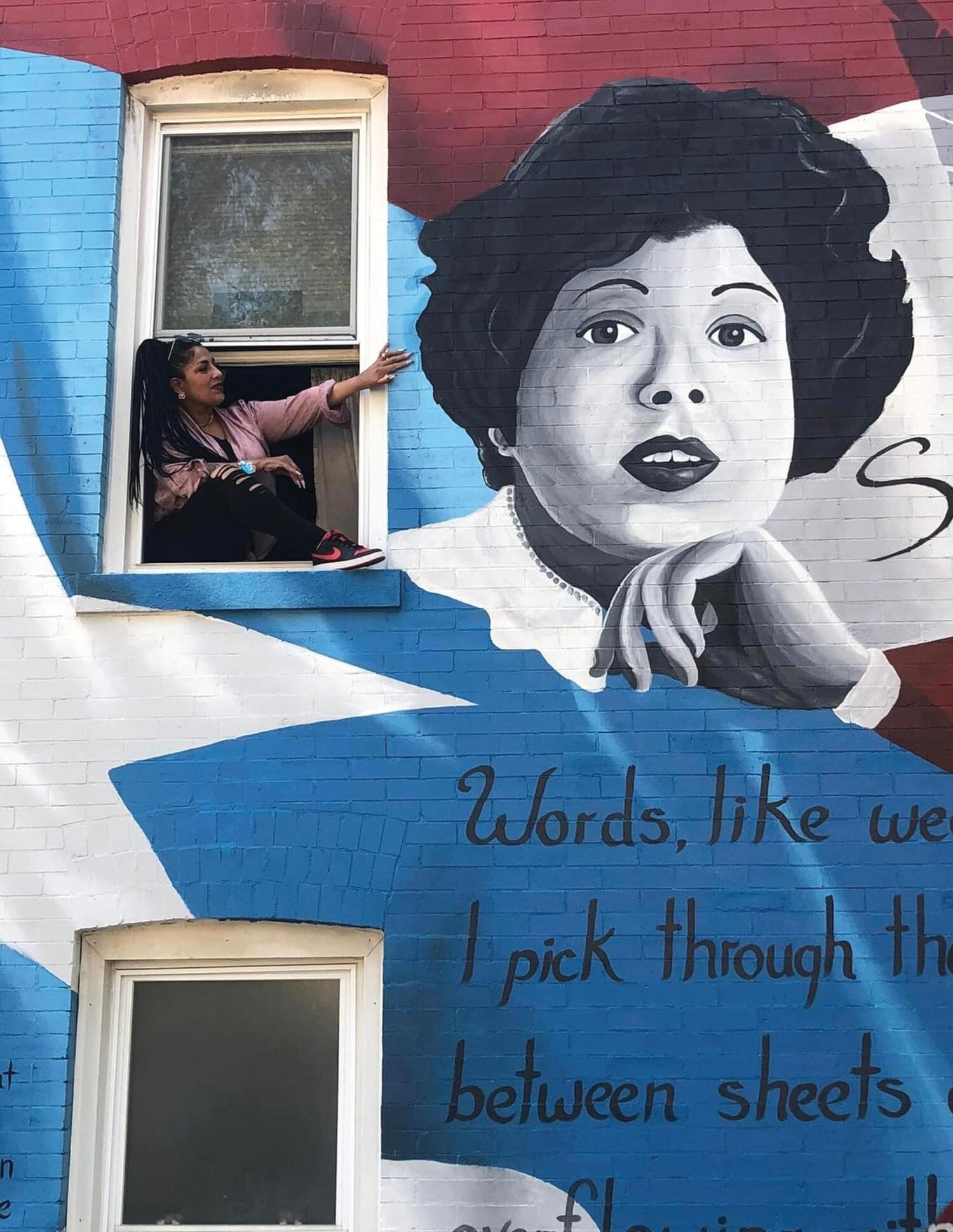 Photograph of a woman with long dark hair and Nike high tops leaning out a window to admire a mural depicting Salima Rivera on top of a Puerto Rican flag