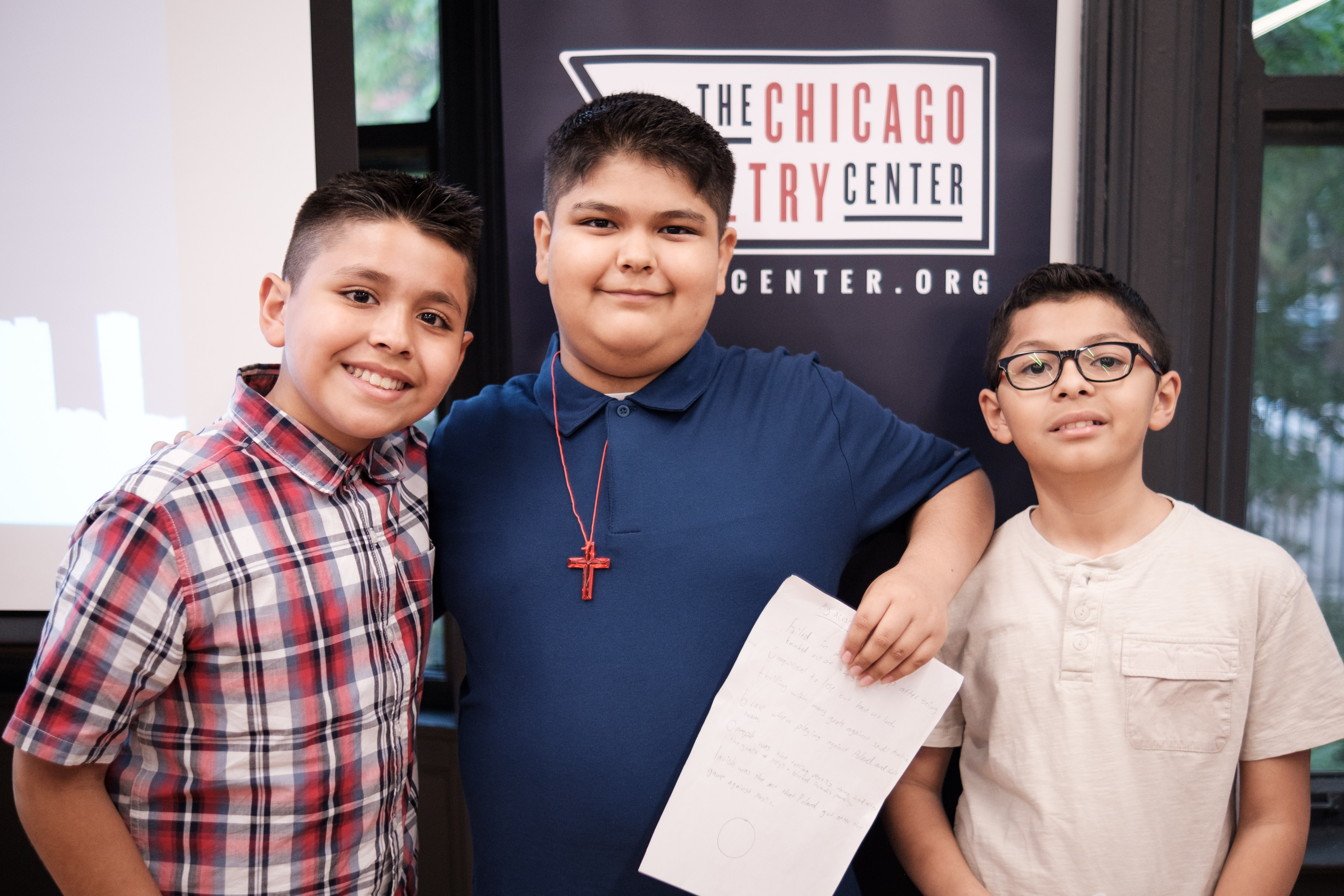 Three smiling young students stand in front of a Chicago Poetry Center sign at the All Schools Reading. One of them holds a piece of paper with their original poem written on it.