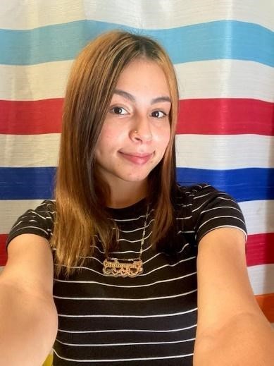 Young woman with her arms outstretched as if she's taking a selfie. She's wearing a black and white striped shirt and standing in front of a red, white, blue, and turquoise cloth.