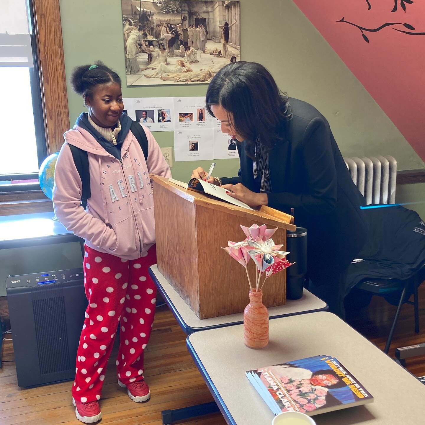 A young woman standing in a classroom setting smiling as another woman, bent over a podium, signs something for her.