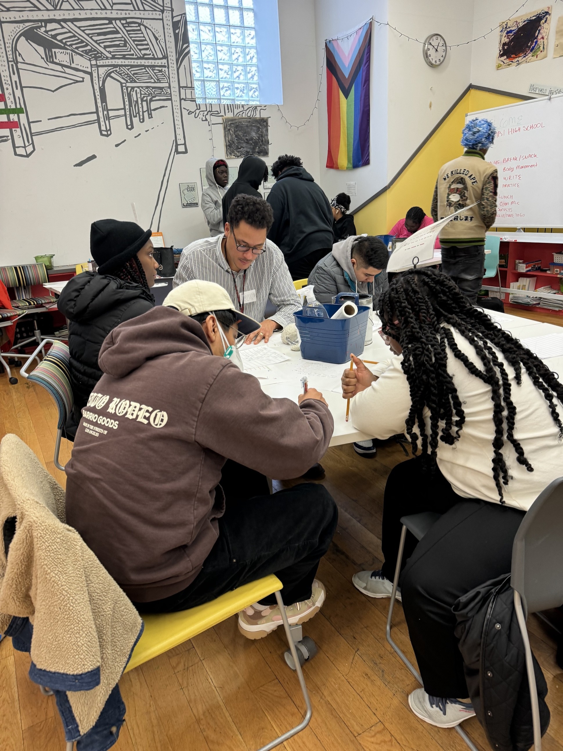 Teens writing at a table in a classroom setting with a rainbow Pride flag in the background.