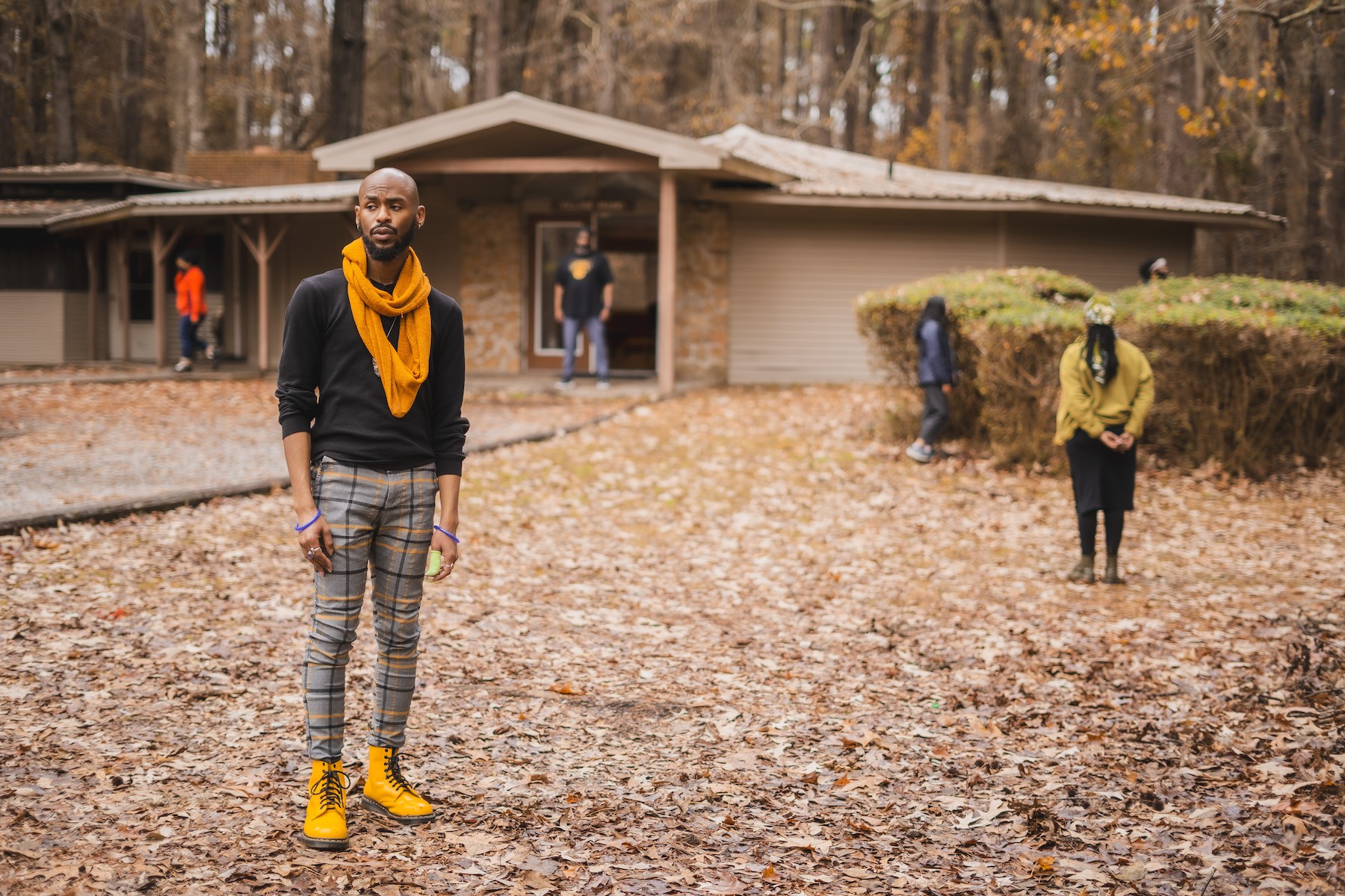  Five adults standing in various places in front of a brown house in a yard covered with brown leaves