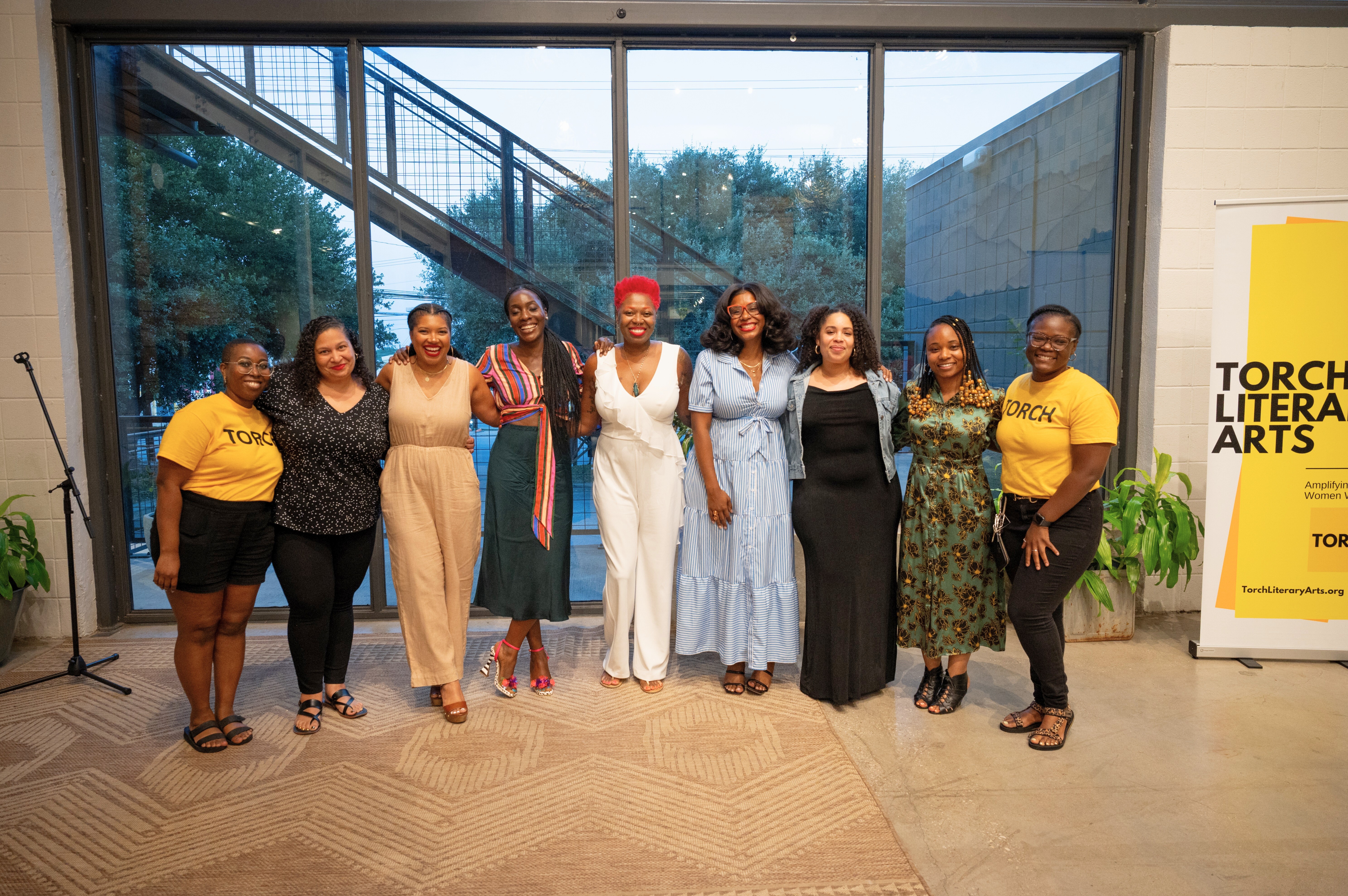 Nine adult women standing linking arms in front of a set of windows with a staircase behind it. Two wear yellow shirts with TORCH in black letters. A banner appears on the left in yellow, white, and black with Torch Literary Arts in black.