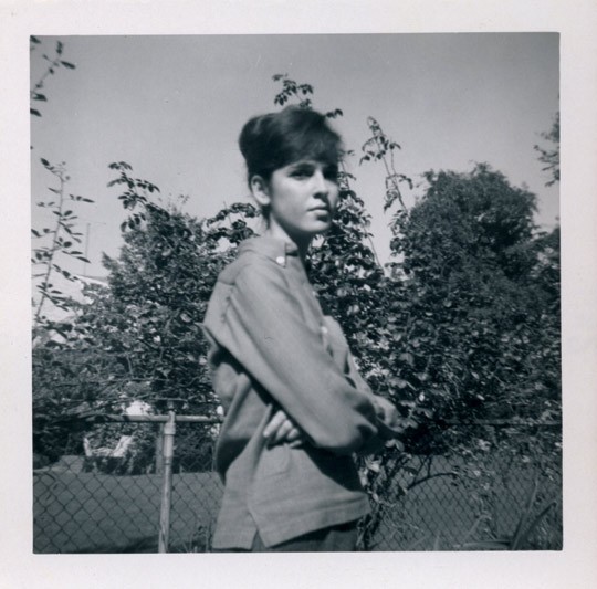 Black and white polaroid of Louise Glück in a jacket in front of a chain-link fence and trees