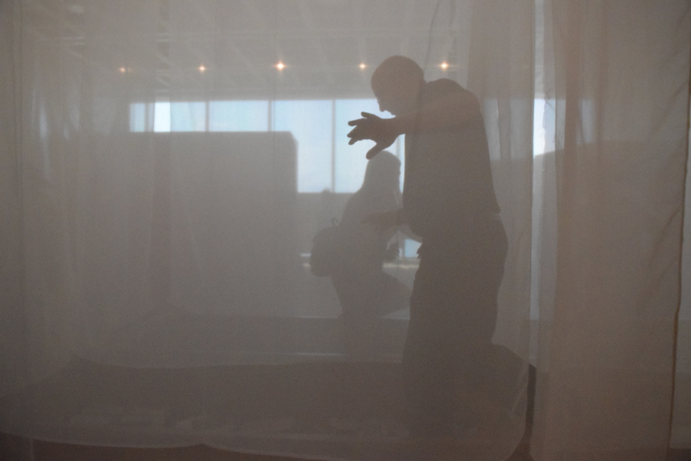 Photo of a man behind translucent screen with woman passing behind him.