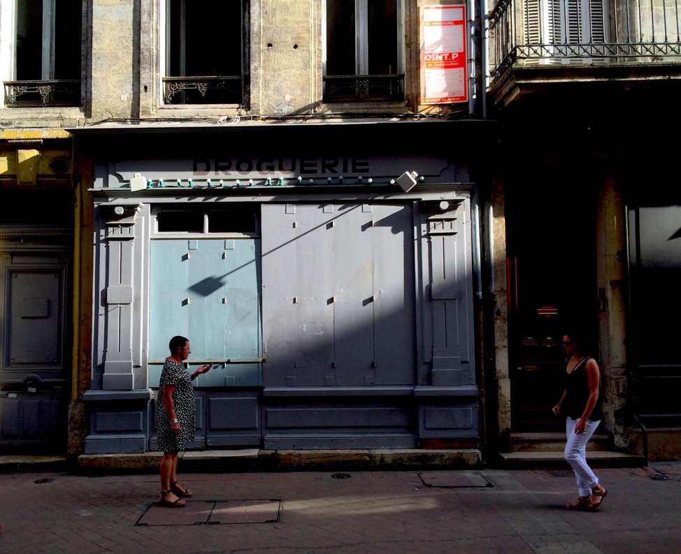 Street photo of two women passing or greeting each other in front of boarded up windows.