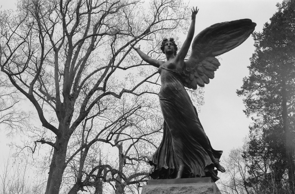 Black and white photo of female angel in Brooklyn cemetary, leaveless trees and sky in background.