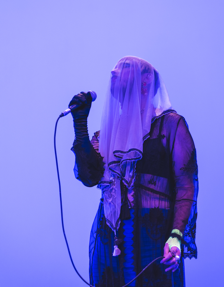 Photo of singer holding mike, wearing a sheer white veil, sheer black top, red earrings. 
