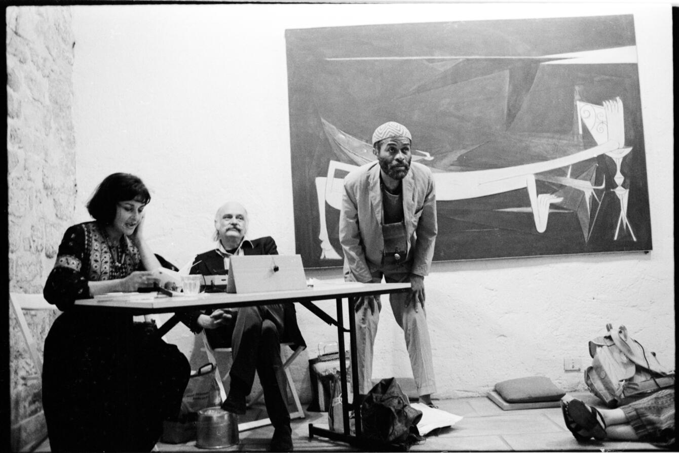 Joyce Mansour sits reading or talking at a table next to a semi-reclined Nanos Valaoritis. Ted Joans stands beside them, leaning over with hands on knees. 