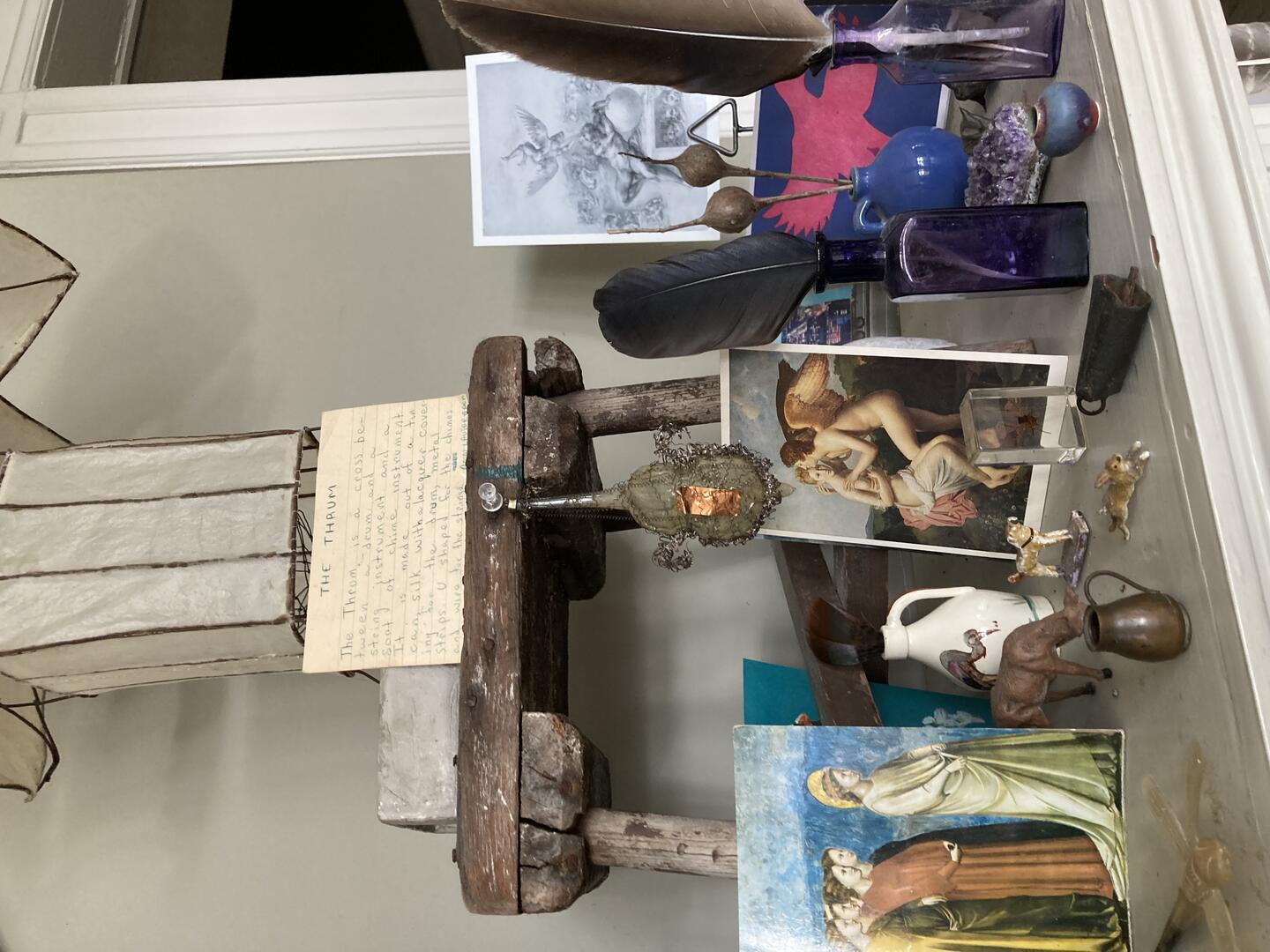 Color photo: Various items on a mantle including art prints, feathers, and figurines